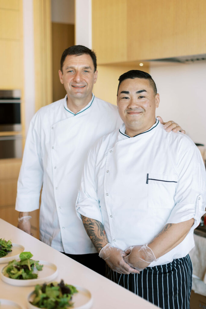 Malibu-Estate-Wedding_Malibu-Wedding_Southern-California-Wedding-Photographer_Lisa-Leanne-Photography_60-683x1024 Two chefs wearing white uniforms and plastic gloves stand side by side in a modern kitchen, smiling at the camera. Plates with fresh green salads are arranged on the counter in front of them. Copyright - Lisa Leanne Photography, California