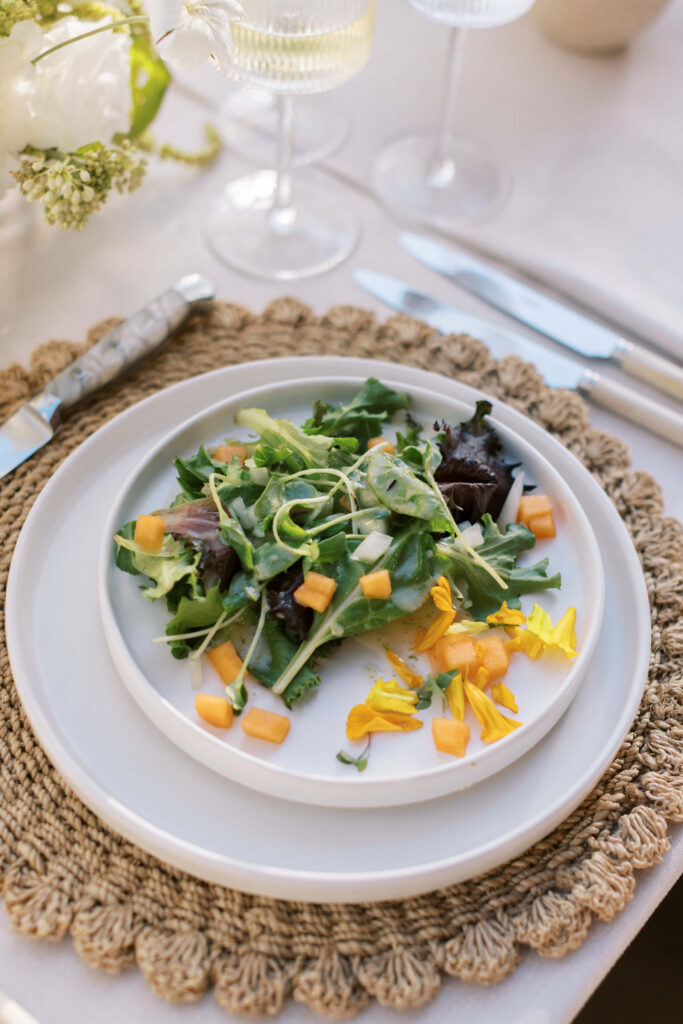Malibu-Estate-Wedding_Malibu-Wedding_Southern-California-Wedding-Photographer_Lisa-Leanne-Photography_61-683x1024 A plate of mixed green salad topped with yellow flower petals and small orange cubes, set on a woven placemat with cutlery and wine glasses nearby. Copyright - Lisa Leanne Photography, California