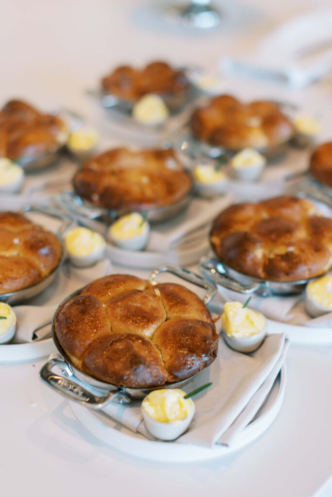 Malibu-Estate-Wedding_Malibu-Wedding_Southern-California-Wedding-Photographer_Lisa-Leanne-Photography_62-683x1024 Small golden-brown bread loaves served in individual round pans, each accompanied by three swirls of butter on a white napkin on a white plate, arranged neatly on a white table. Copyright - Lisa Leanne Photography, California