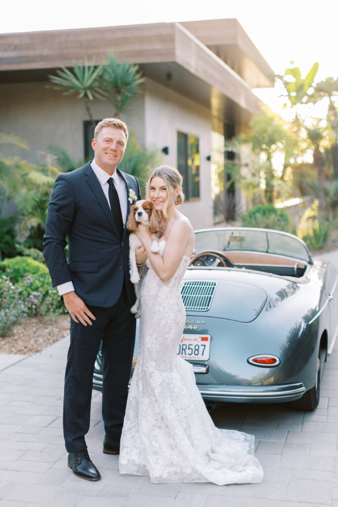 Malibu-Estate-Wedding_Malibu-Wedding_Southern-California-Wedding-Photographer_Lisa-Leanne-Photography_64-683x1024 A bride in a lace dress and a groom in a dark suit stand smiling beside a vintage convertible car. The bride holds a small brown and white dog. Modern house and lush greenery are visible in the background. Copyright - Lisa Leanne Photography, California