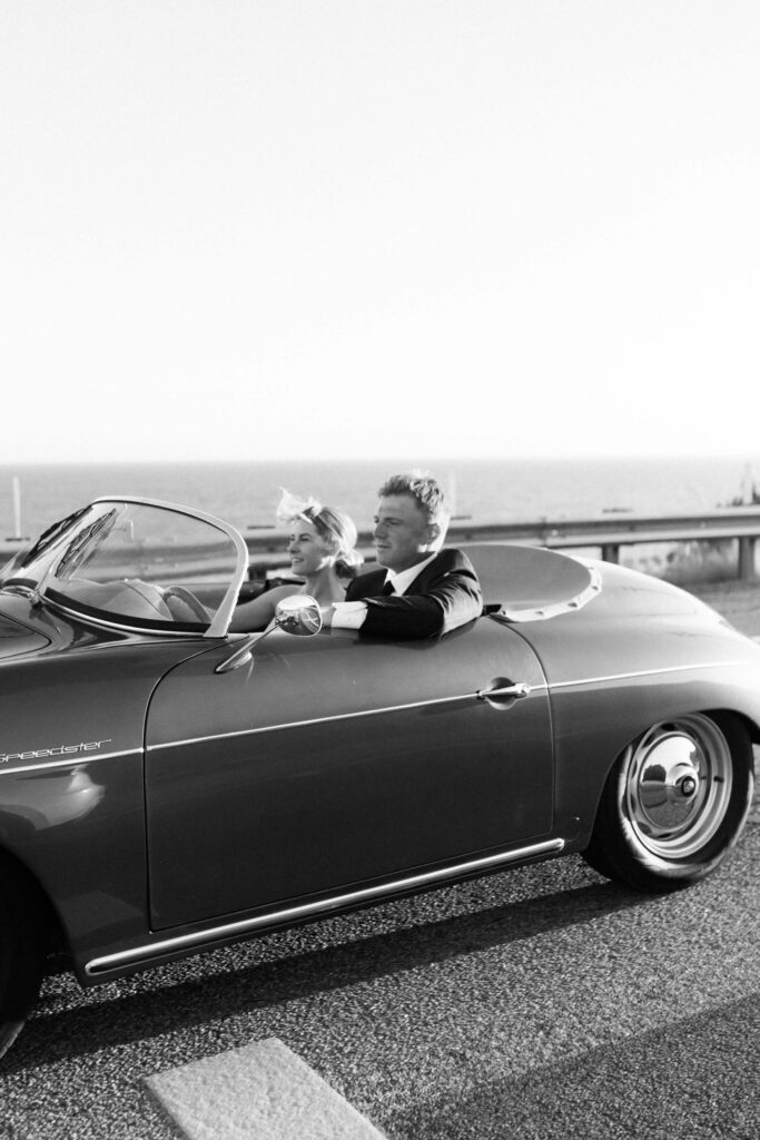 Malibu-Estate-Wedding_Malibu-Wedding_Southern-California-Wedding-Photographer_Lisa-Leanne-Photography_66-683x1024 A bride and groom in formal attire drive a vintage convertible car along a coastal road, smiling with the ocean visible in the background. The image is in black and white. Copyright - Lisa Leanne Photography, California
