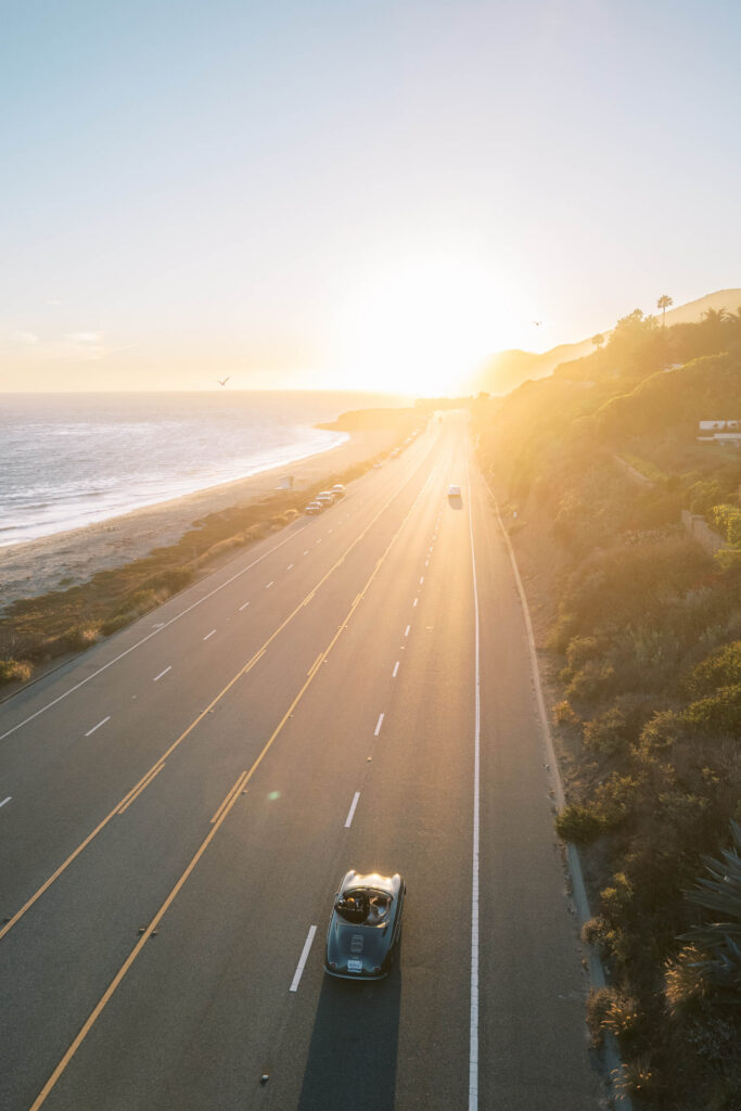 Malibu-Estate-Wedding_Malibu-Wedding_Southern-California-Wedding-Photographer_Lisa-Leanne-Photography_68-683x1024 A convertible car drives along a coastal highway at sunset, with the sun low on the horizon, casting long shadows over the road; the ocean is on the left and green hills are on the right. Copyright - Lisa Leanne Photography, California