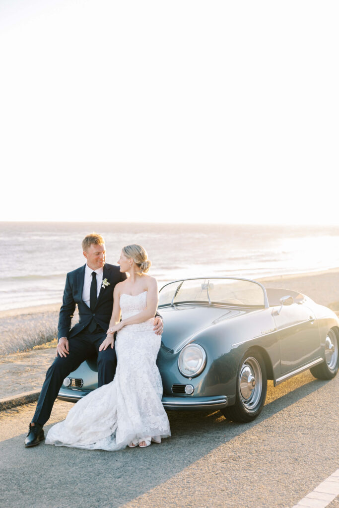 Malibu-Estate-Wedding_Malibu-Wedding_Southern-California-Wedding-Photographer_Lisa-Leanne-Photography_69-683x1024 A bride in a white wedding dress and a groom in a dark suit sit together on the hood of a vintage car by the ocean at sunset, sharing an intimate moment. Copyright - Lisa Leanne Photography, California