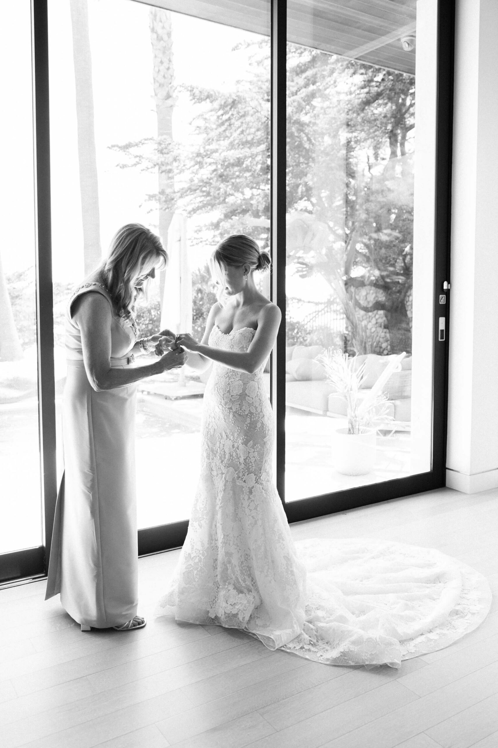 A bride in a lace wedding gown stands by large windows as another woman, possibly her mother, helps her with jewelry. Natural light fills the room, highlighting their elegant dresses and the bride’s train. Copyright - Lisa Leanne Photography, California