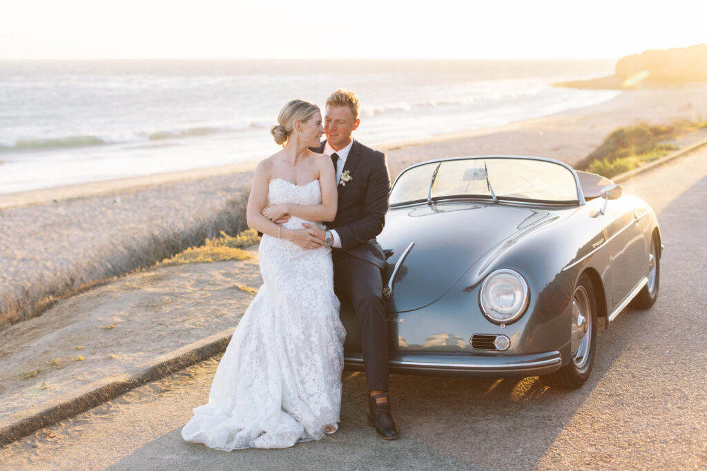 A bride in a white gown and a groom in a dark suit sit together on the hood of a vintage car by the beach at sunset, embracing and smiling. Copyright - Lisa Leanne Photography, California
