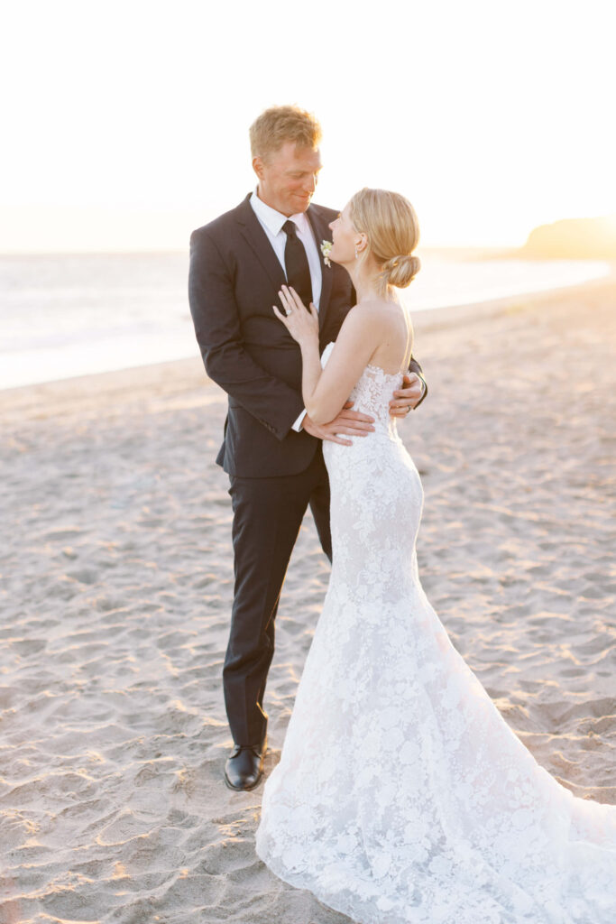 Malibu-Estate-Wedding_Malibu-Wedding_Southern-California-Wedding-Photographer_Lisa-Leanne-Photography_71-683x1024 A bride in a lace wedding dress and a groom in a suit stand on a sandy beach at sunset, embracing and smiling at each other. Copyright - Lisa Leanne Photography, California