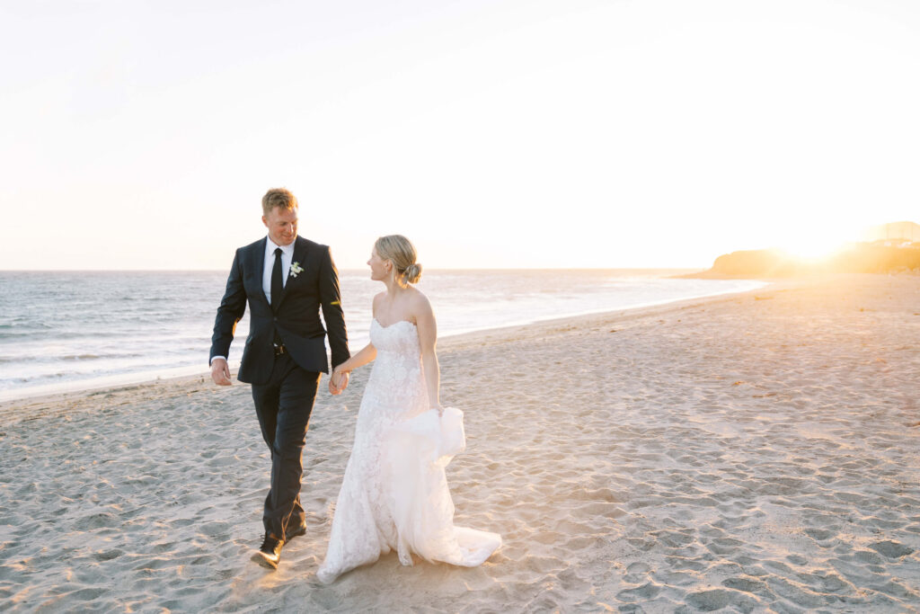 Malibu-Estate-Wedding_Malibu-Wedding_Southern-California-Wedding-Photographer_Lisa-Leanne-Photography_73-1024x683 A bride in a white gown and a groom in a dark suit walk hand-in-hand along a sandy beach at sunset, with gentle waves and golden light in the background. Copyright - Lisa Leanne Photography, California