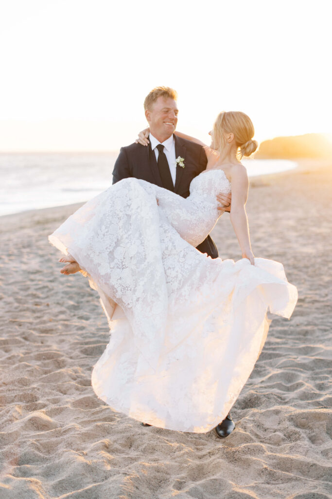 Malibu-Estate-Wedding_Malibu-Wedding_Southern-California-Wedding-Photographer_Lisa-Leanne-Photography_74-683x1024 A groom in a black suit carries his bride, who wears a white lace wedding dress, along a sandy beach at sunset. Both are smiling and looking at each other, bathed in warm, golden light. Copyright - Lisa Leanne Photography, California