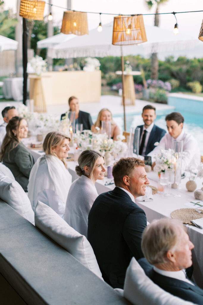 A group of people dressed formally sit around a long, elegantly decorated table by a pool, enjoying an outdoor event with white flowers, candles, and string lights in the background. Copyright – Lisa Leanne Photography, California A group of people dressed formally sit around a long, elegantly decorated table by a pool, enjoying an outdoor event with white flowers, candles, and string lights in the background. Copyright - Lisa Leanne Photography, California