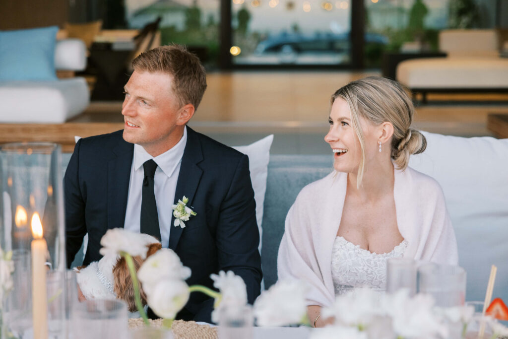 A bride and groom sit side by side at a decorated table, smiling and looking to their left. The bride wears a white dress and the groom is in a dark suit with a boutonniere. White flowers and candles decorate the table. Copyright – Lisa Leanne Photography, California A bride and groom sit side by side at a decorated table, smiling and looking to their left. The bride wears a white dress and the groom is in a dark suit with a boutonniere. White flowers and candles decorate the table. Copyright - Lisa Leanne Photography, California