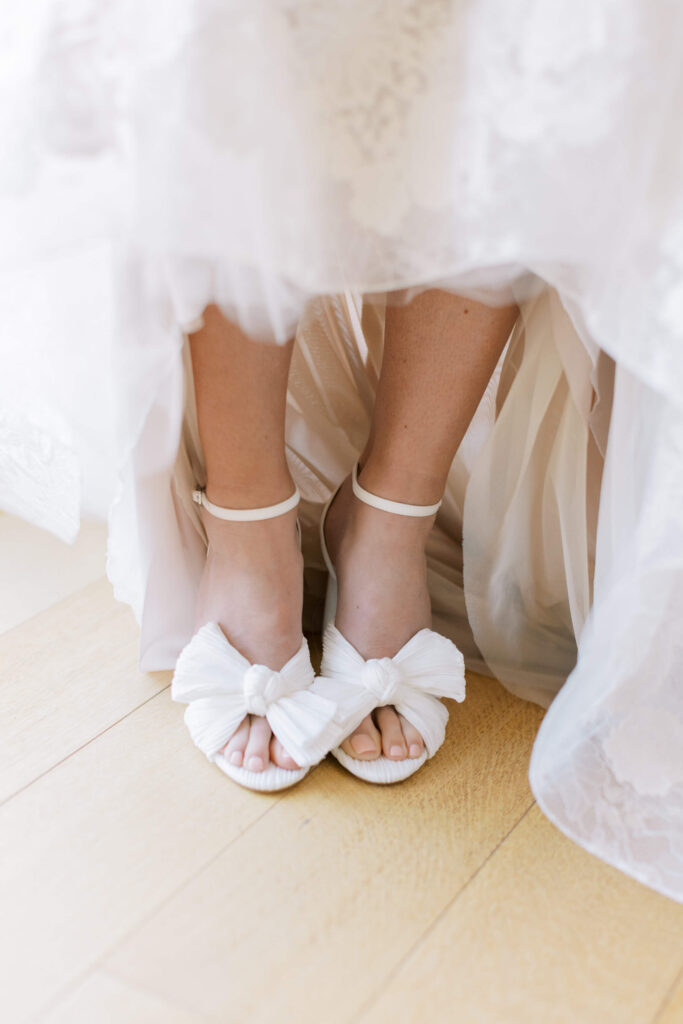 Malibu-Estate-Wedding_Malibu-Wedding_Southern-California-Wedding-Photographer_Lisa-Leanne-Photography_8-683x1024 A close-up of a person in a white wedding dress, showing their feet in white, open-toed heels with ankle straps and large bows, standing on a wooden floor. Copyright - Lisa Leanne Photography, California