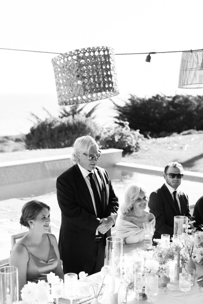 A man in a suit stands giving a speech at an outdoor table set for a formal event. Three seated guests, dressed elegantly, listen and smile. Decorative lanterns hang above, with greenery and water in the background. Copyright – Lisa Leanne Photography, California A man in a suit stands giving a speech at an outdoor table set for a formal event. Three seated guests, dressed elegantly, listen and smile. Decorative lanterns hang above, with greenery and water in the background. Copyright - Lisa Leanne Photography, California