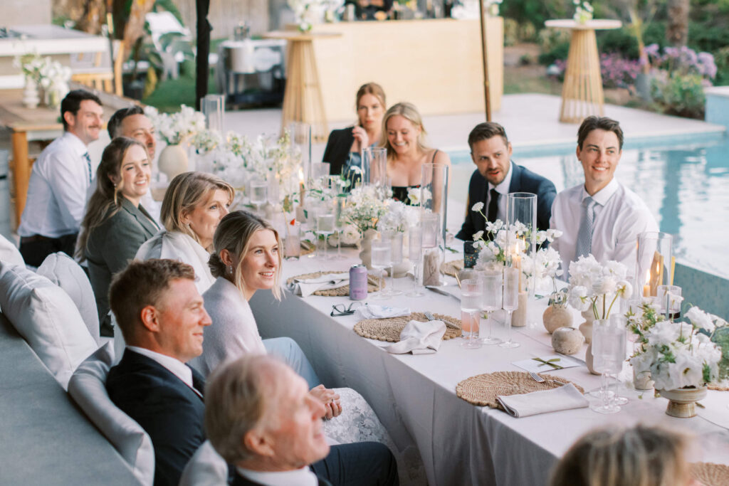 A group of people dressed formally sit around a long, elegant outdoor dining table decorated with white flowers and candles, smiling and enjoying a celebration by a pool. Copyright – Lisa Leanne Photography, California A group of people dressed formally sit around a long, elegant outdoor dining table decorated with white flowers and candles, smiling and enjoying a celebration by a pool. Copyright - Lisa Leanne Photography, California