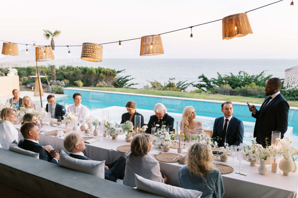 A group of people in formal attire sit at a long outdoor table decorated with white flowers, dining near a pool with an ocean view, while a man stands and gives a speech. Hanging lights and woven lamps add a warm ambiance. Copyright – Lisa Leanne Photography, California A group of people in formal attire sit at a long outdoor table decorated with white flowers, dining near a pool with an ocean view, while a man stands and gives a speech. Hanging lights and woven lamps add a warm ambiance. Copyright - Lisa Leanne Photography, California