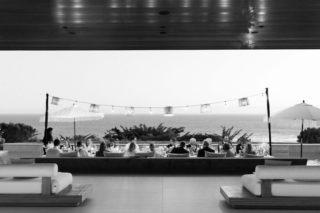 Malibu-Estate-Wedding_Malibu-Wedding_Southern-California-Wedding-Photographer_Lisa-Leanne-Photography_85-1024x683 A group of people dine outdoors under string lights, overlooking the ocean. The scene is framed by modern lounge chairs and an umbrella, with trees and water visible in the background. The image is in black and white. Copyright - Lisa Leanne Photography, California