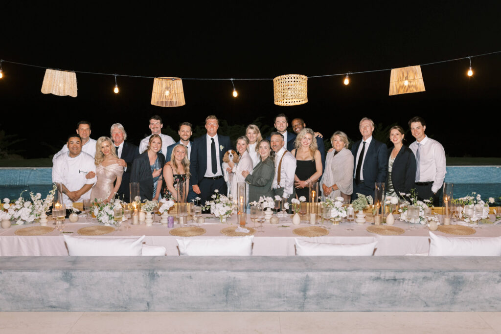 Malibu-Estate-Wedding_Malibu-Wedding_Southern-California-Wedding-Photographer_Lisa-Leanne-Photography_86-1024x683 A group of people dressed in formal attire pose together behind a long, elegantly set outdoor dinner table with floral centerpieces, under string lights and hanging lanterns at night. Copyright - Lisa Leanne Photography, California