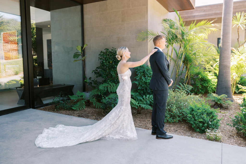 Malibu-Estate-Wedding_Malibu-Wedding_Southern-California-Wedding-Photographer_Lisa-Leanne-Photography_9-1024x683 A bride in a white lace gown touches the shoulder of a groom in a black suit during a first look outside a modern building, surrounded by greenery and palm trees. Copyright - Lisa Leanne Photography, California