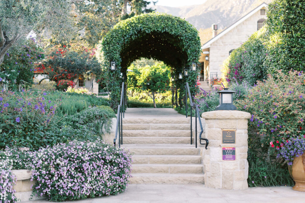 A stone staircase leads up to an arched trellis covered in greenery and string lights, surrounded by lush gardens and colorful flowers, with a stone building in the background. Copyright - Lisa Leanne Photography, California