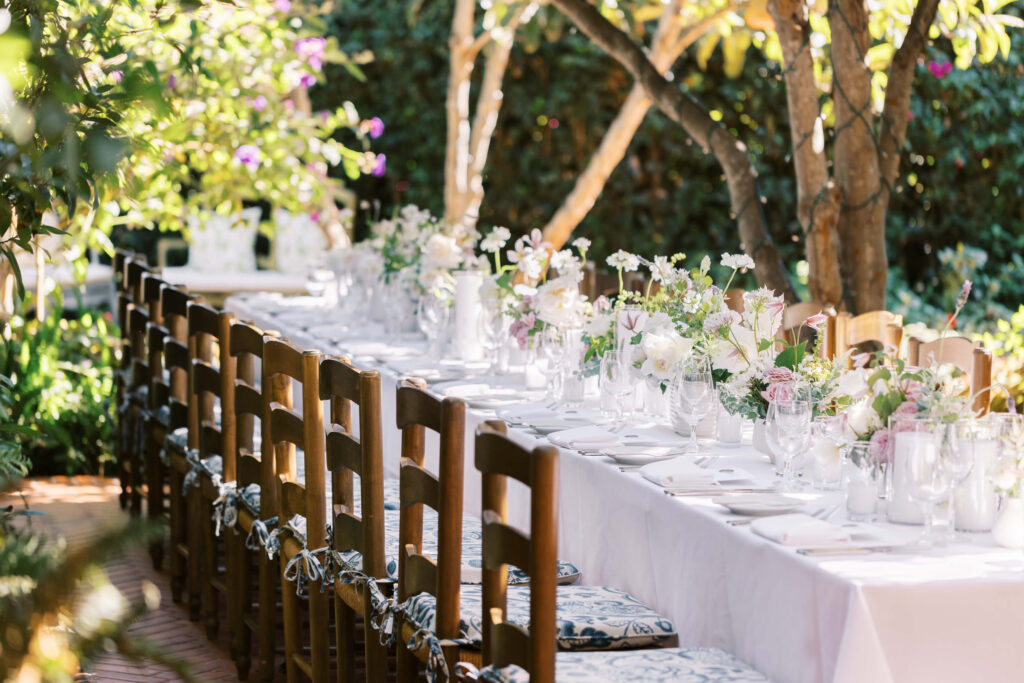 San-Ysidro-Ranch-Rehearsal-Dinner_Santa-Barbara-Wedding_Southern-California-Wedding-Photographer_Lisa-Leanne-Photography_11-1024x683 A long outdoor dining table with white tablecloth, elegant glassware, and floral centerpieces is surrounded by wooden chairs in a sunlit garden setting. Copyright - Lisa Leanne Photography, California