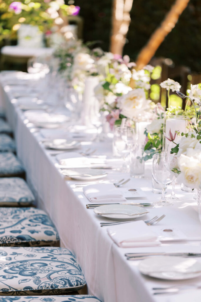 San-Ysidro-Ranch-Rehearsal-Dinner_Santa-Barbara-Wedding_Southern-California-Wedding-Photographer_Lisa-Leanne-Photography_12-683x1024 A long, outdoor table set for a formal event with white tablecloth, floral centerpieces, glassware, plates, and cutlery. Cushioned chairs with blue and white patterns are arranged along the table. Copyright - Lisa Leanne Photography, California