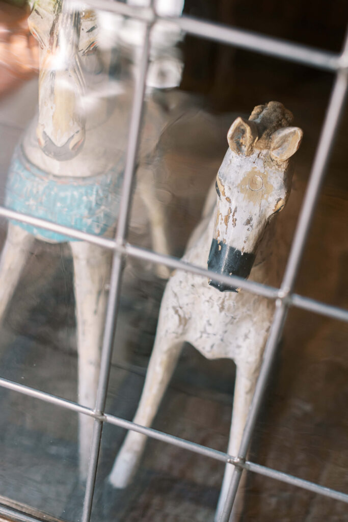 San-Ysidro-Ranch-Rehearsal-Dinner_Santa-Barbara-Wedding_Southern-California-Wedding-Photographer_Lisa-Leanne-Photography_13-683x1024 Two vintage, weathered wooden horse figurines are seen through a window with square metal grilles. The paint on the horses is chipped, adding to their rustic, antique appearance. Copyright - Lisa Leanne Photography, California