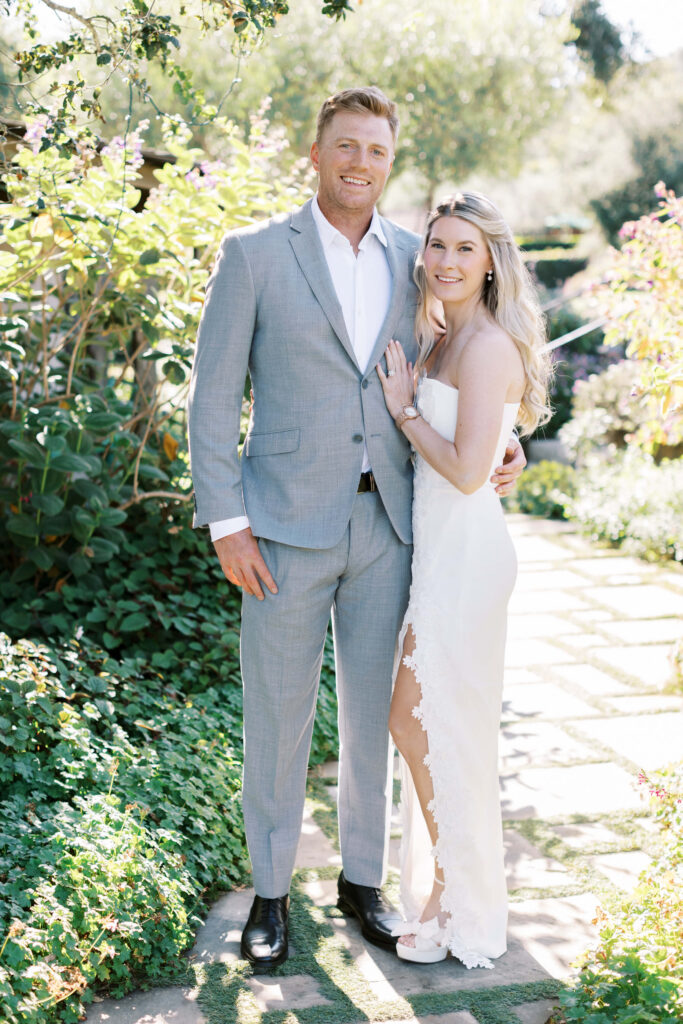 San-Ysidro-Ranch-Rehearsal-Dinner_Santa-Barbara-Wedding_Southern-California-Wedding-Photographer_Lisa-Leanne-Photography_14-683x1024 A smiling couple poses outdoors on a stone path surrounded by lush greenery. The man wears a light gray suit and white shirt; the woman wears a strapless white dress with a lace slit. Both look happy and relaxed in the sunlight. Copyright - Lisa Leanne Photography, California