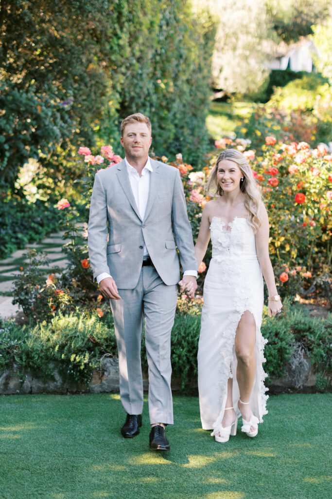 San-Ysidro-Ranch-Rehearsal-Dinner_Santa-Barbara-Wedding_Southern-California-Wedding-Photographer_Lisa-Leanne-Photography_17-683x1024 A bride in a white dress with floral details and a groom in a light gray suit walk hand in hand outdoors, surrounded by blooming flowers and greenery. Copyright - Lisa Leanne Photography, California