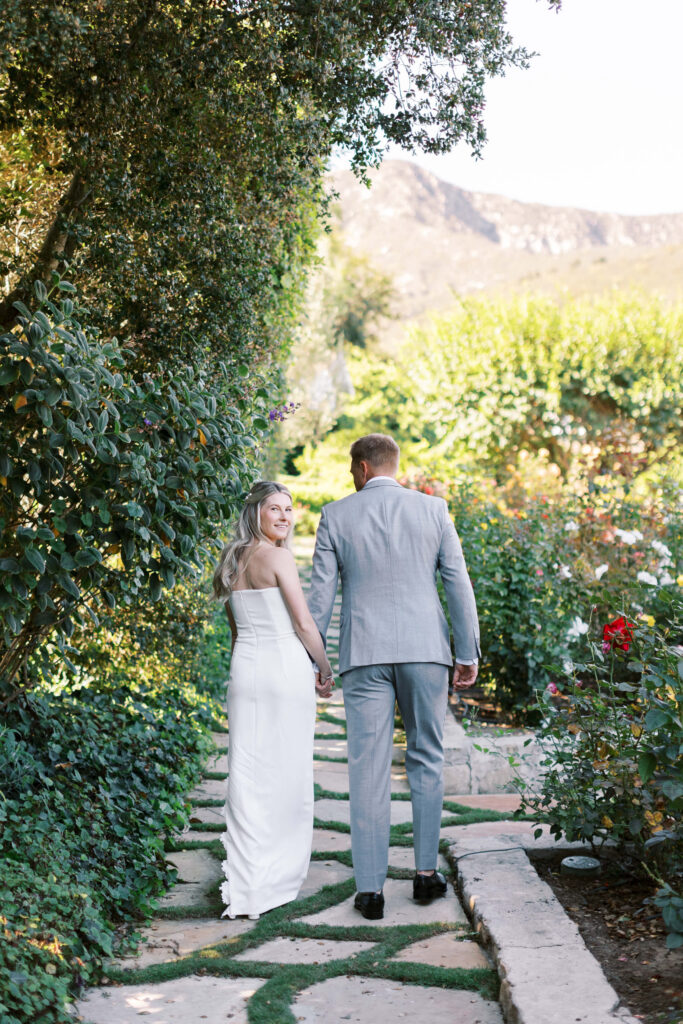 San-Ysidro-Ranch-Rehearsal-Dinner_Santa-Barbara-Wedding_Southern-California-Wedding-Photographer_Lisa-Leanne-Photography_18-683x1024 A couple, dressed in wedding attire, walks hand-in-hand down a stone path surrounded by lush greenery and flowers. The woman looks back and smiles, while the man faces forward. Mountains are visible in the background. Copyright - Lisa Leanne Photography, California