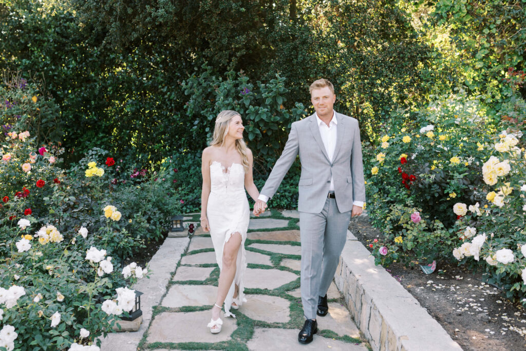San-Ysidro-Ranch-Rehearsal-Dinner_Santa-Barbara-Wedding_Southern-California-Wedding-Photographer_Lisa-Leanne-Photography_19-1024x683 A couple walks hand in hand along a stone path in a garden filled with blooming flowers. The woman wears a white dress and the man wears a light gray suit. Lush greenery and colorful roses surround them. Copyright - Lisa Leanne Photography, California