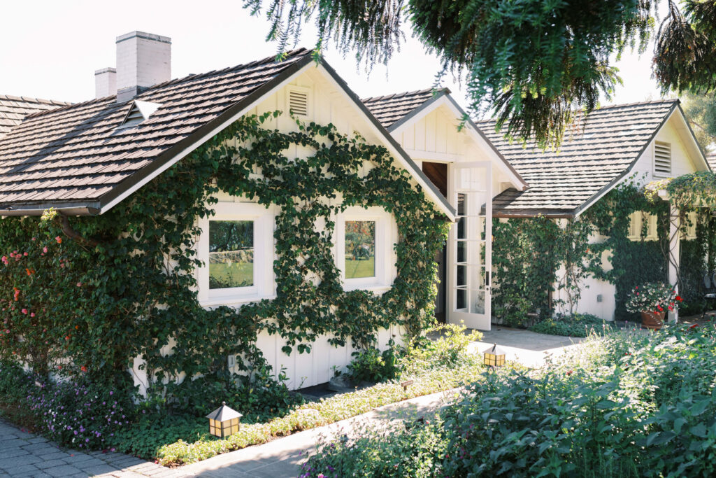 San-Ysidro-Ranch-Rehearsal-Dinner_Santa-Barbara-Wedding_Southern-California-Wedding-Photographer_Lisa-Leanne-Photography_2-1024x683 A charming white cottage with a shingled roof is covered in green ivy and surrounded by lush plants and flowers. Sunlight illuminates the garden and pathway leading to the glass front door. Copyright - Lisa Leanne Photography, California