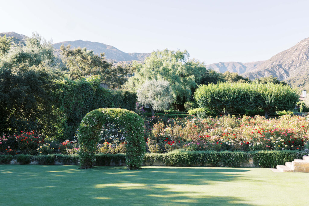 San-Ysidro-Ranch-Rehearsal-Dinner_Santa-Barbara-Wedding_Southern-California-Wedding-Photographer_Lisa-Leanne-Photography_22-1024x683 A lush garden with a green archway, colorful blooming flowers, trimmed hedges, and trees, set against a backdrop of mountains under a clear sky. Copyright - Lisa Leanne Photography, California