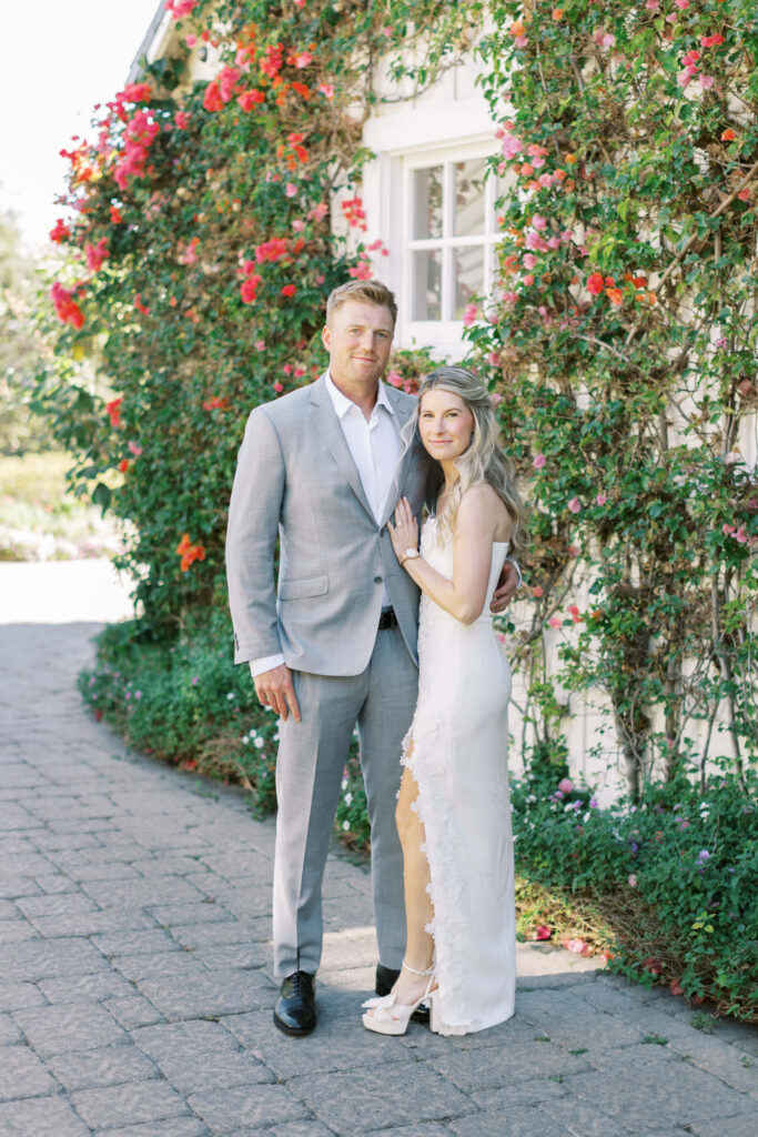 San-Ysidro-Ranch-Rehearsal-Dinner_Santa-Barbara-Wedding_Southern-California-Wedding-Photographer_Lisa-Leanne-Photography_23-683x1024 A man in a light gray suit stands next to a woman in a white dress with floral trim. They are outside on a stone path beside a building covered in green vines and pink flowers, posing for a photo together. Copyright - Lisa Leanne Photography, California