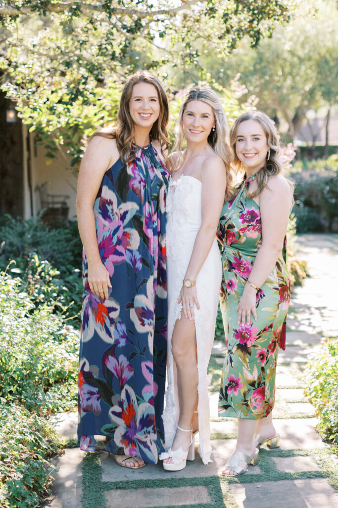 San-Ysidro-Ranch-Rehearsal-Dinner_Santa-Barbara-Wedding_Southern-California-Wedding-Photographer_Lisa-Leanne-Photography_24-683x1024 Three women pose outdoors on a garden path. The woman in the center wears a white dress, while the others wear colorful floral dresses. Sunlight filters through the trees, creating a bright and cheerful atmosphere. Copyright - Lisa Leanne Photography, California