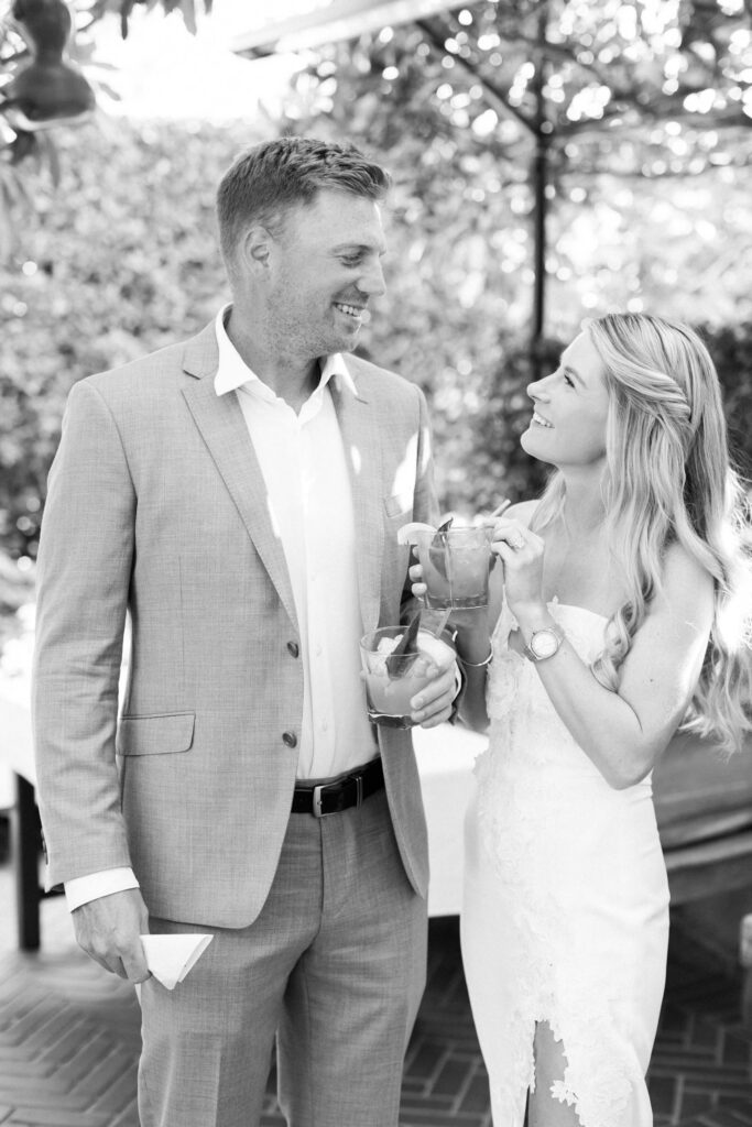 San-Ysidro-Ranch-Rehearsal-Dinner_Santa-Barbara-Wedding_Southern-California-Wedding-Photographer_Lisa-Leanne-Photography_25-683x1024 A man in a suit and a woman in a white dress smile at each other while holding drinks outdoors, surrounded by greenery and sunlight. Copyright - Lisa Leanne Photography, California