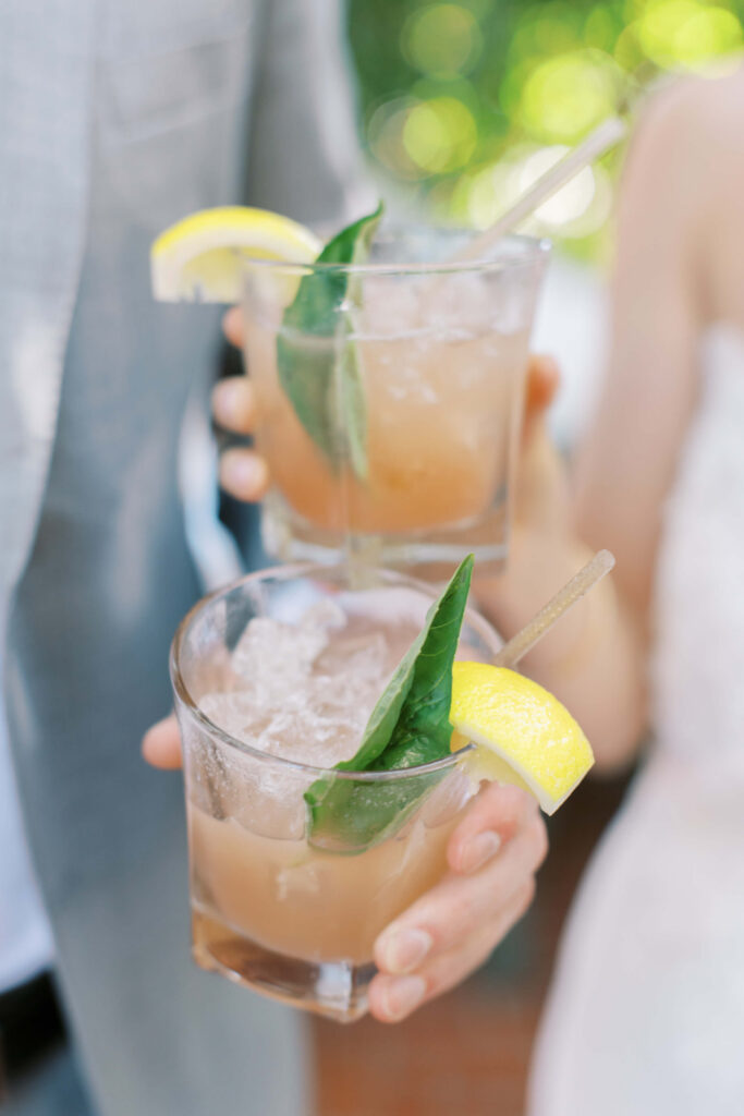 San-Ysidro-Ranch-Rehearsal-Dinner_Santa-Barbara-Wedding_Southern-California-Wedding-Photographer_Lisa-Leanne-Photography_26-683x1024 Two hands holding glasses of a light brown iced cocktail garnished with a fresh basil leaf and a lemon wedge, with a blurred green background. Copyright - Lisa Leanne Photography, California