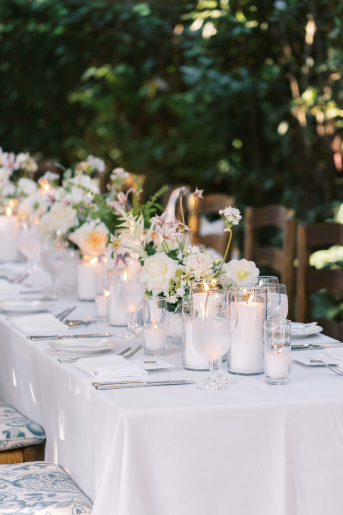 San-Ysidro-Ranch-Rehearsal-Dinner_Santa-Barbara-Wedding_Southern-California-Wedding-Photographer_Lisa-Leanne-Photography_27-683x1024 A beautifully set outdoor table with a white tablecloth, white flowers in vases, and lit candles arranged along the center; place settings with glasses, plates, and silverware are neatly arranged. Lush greenery is in the background. Copyright - Lisa Leanne Photography, California
