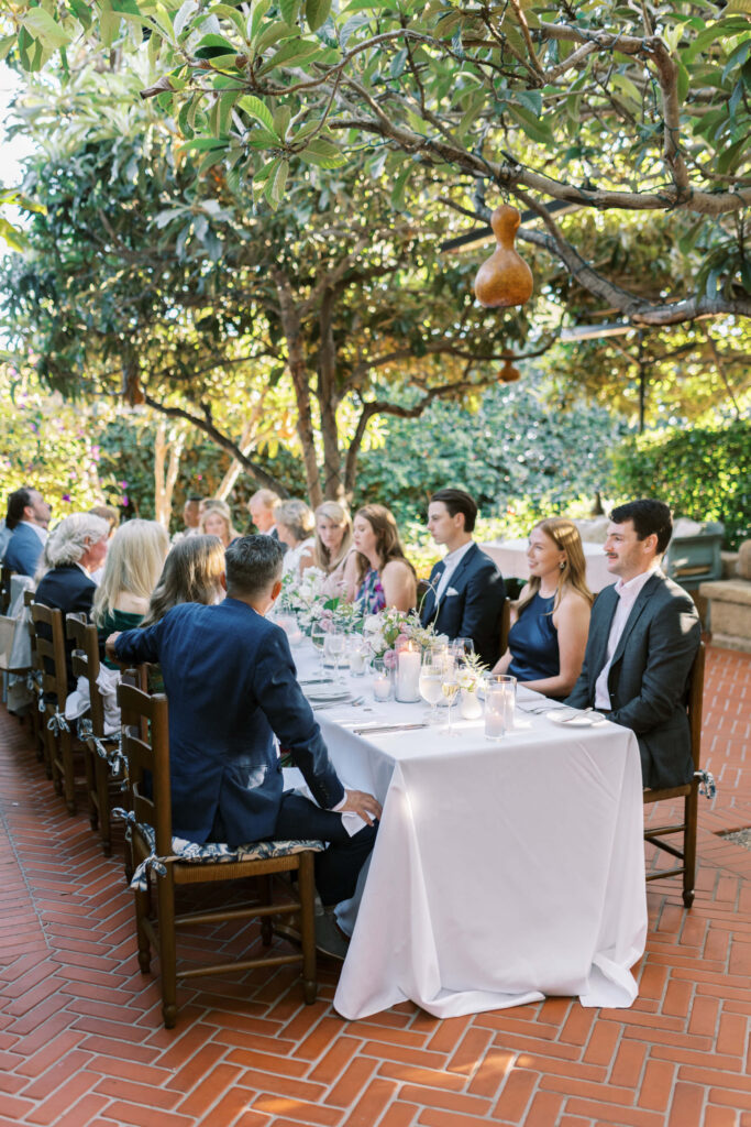 San-Ysidro-Ranch-Rehearsal-Dinner_Santa-Barbara-Wedding_Southern-California-Wedding-Photographer_Lisa-Leanne-Photography_29-683x1024 A group of people dressed formally sit around a long, white-clothed table outdoors under leafy trees, enjoying a meal together in a sunlit garden setting. Copyright - Lisa Leanne Photography, California