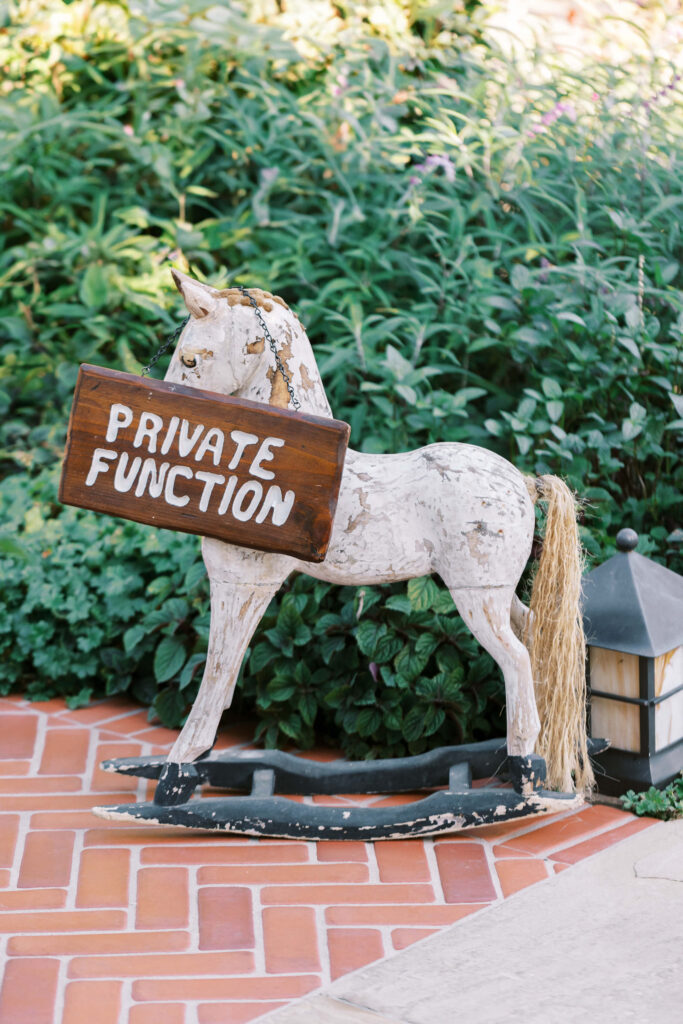 San-Ysidro-Ranch-Rehearsal-Dinner_Santa-Barbara-Wedding_Southern-California-Wedding-Photographer_Lisa-Leanne-Photography_3-683x1024 A weathered white rocking horse stands on a brick patio, holding a wooden sign that reads "PRIVATE FUNCTION" in white letters. Lush green plants and a lantern are in the background. Copyright - Lisa Leanne Photography, California