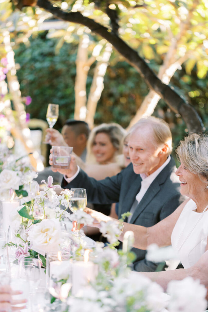 San-Ysidro-Ranch-Rehearsal-Dinner_Santa-Barbara-Wedding_Southern-California-Wedding-Photographer_Lisa-Leanne-Photography_30-683x1024 A group of people sit at a beautifully decorated outdoor table with white flowers, raising glasses in a toast, smiling and enjoying a joyful celebration under dappled sunlight. Copyright - Lisa Leanne Photography, California