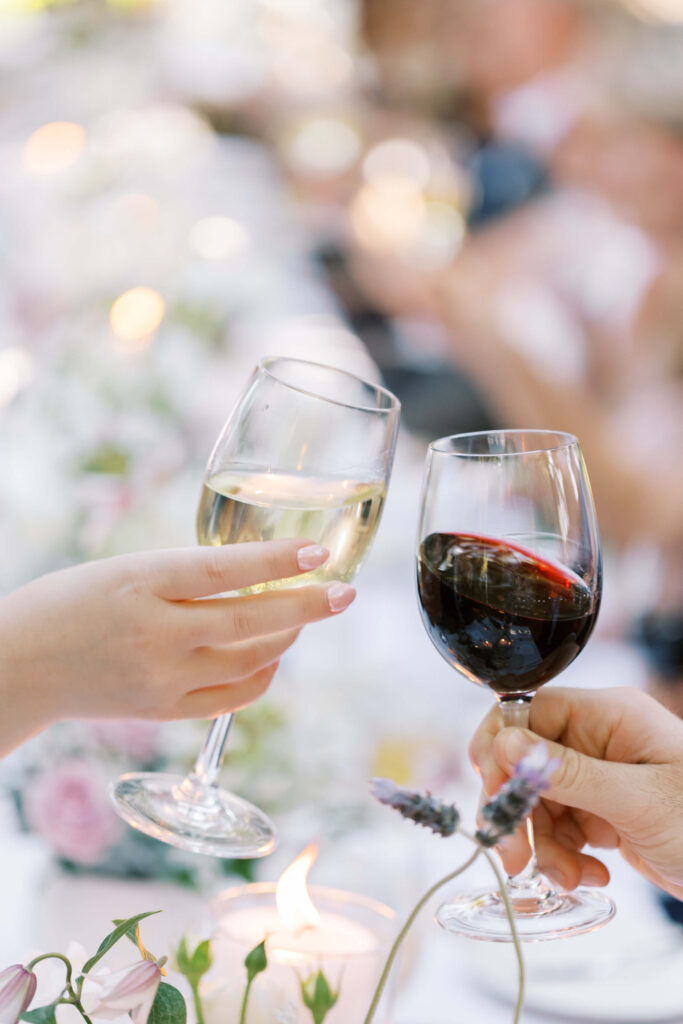 San-Ysidro-Ranch-Rehearsal-Dinner_Santa-Barbara-Wedding_Southern-California-Wedding-Photographer_Lisa-Leanne-Photography_31-683x1024 Two people clinking glasses, one holding a glass of white wine and the other a glass of red wine, with a blurred, elegant background and flowers on the table. Copyright - Lisa Leanne Photography, California