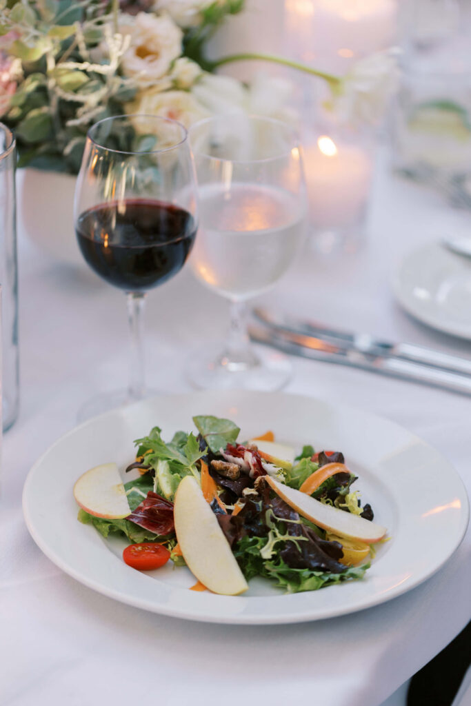 San-Ysidro-Ranch-Rehearsal-Dinner_Santa-Barbara-Wedding_Southern-California-Wedding-Photographer_Lisa-Leanne-Photography_33-683x1024 A plate of mixed green salad with apple slices, cherry tomatoes, and nuts sits on a white tablecloth beside a glass of red wine, a glass of water, and silver cutlery, with candles and flowers in the background. Copyright - Lisa Leanne Photography, California