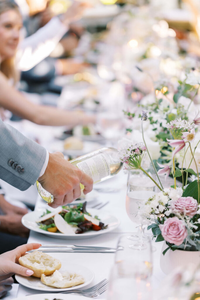 San-Ysidro-Ranch-Rehearsal-Dinner_Santa-Barbara-Wedding_Southern-California-Wedding-Photographer_Lisa-Leanne-Photography_34-683x1024 A person in a light gray suit pours white wine into a glass at a beautifully decorated table with floral arrangements, salads, and plates, surrounded by people enjoying a meal together. Copyright - Lisa Leanne Photography, California