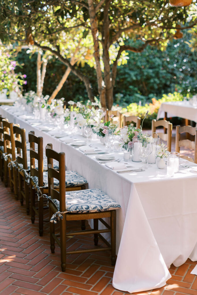 San-Ysidro-Ranch-Rehearsal-Dinner_Santa-Barbara-Wedding_Southern-California-Wedding-Photographer_Lisa-Leanne-Photography_6-683x1024 A long outdoor dining table with a white tablecloth, set with glassware, plates, and small floral centerpieces. Wooden chairs with patterned cushions line both sides, surrounded by greenery and dappled sunlight. Copyright - Lisa Leanne Photography, California
