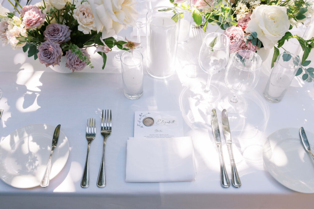 San-Ysidro-Ranch-Rehearsal-Dinner_Santa-Barbara-Wedding_Southern-California-Wedding-Photographer_Lisa-Leanne-Photography_7-1024x683 An elegant table setting with plates, cutlery, wine glasses, and a folded napkin on a white tablecloth. A floral centerpiece with white and pink flowers decorates the table, with sunlight casting soft shadows. Copyright - Lisa Leanne Photography, California