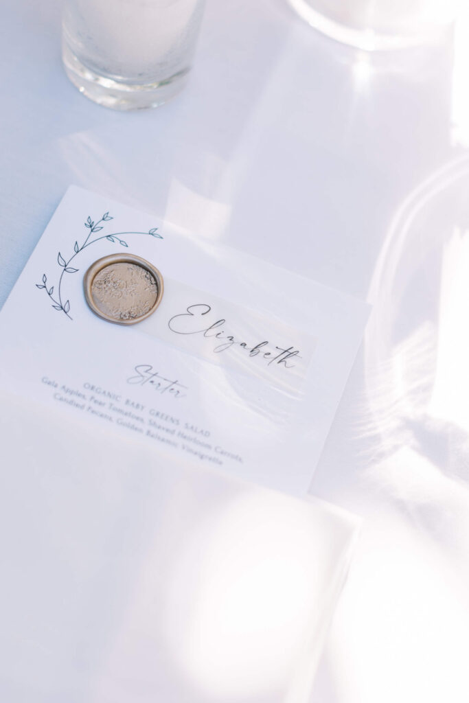 San-Ysidro-Ranch-Rehearsal-Dinner_Santa-Barbara-Wedding_Southern-California-Wedding-Photographer_Lisa-Leanne-Photography_9-683x1024 A place card with the name "Elizabeth" written in elegant script, decorated with a sprig illustration and topped with a coin, rests on a white tablecloth with a glass candle nearby. Sunlight creates soft shadows. Copyright - Lisa Leanne Photography, California
