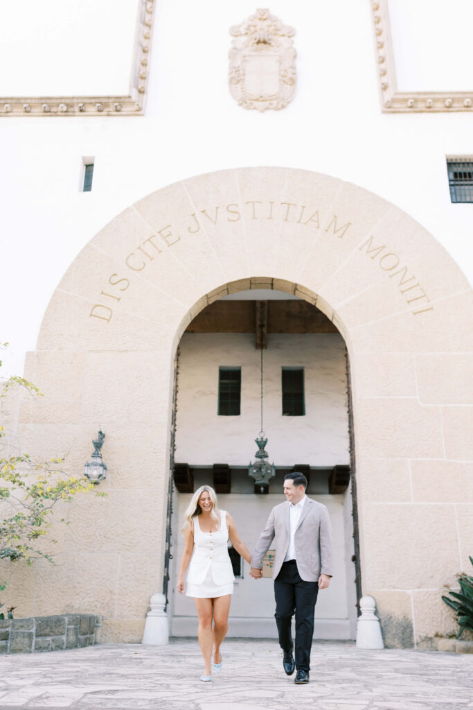 A woman in a white dress and a man in a tan jacket hold hands while walking down steps beneath a large stone arch that reads “Discite Justitiam Moniti.”. Copyright - Lisa Leanne Photography, California