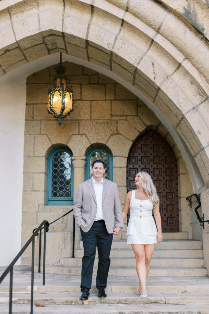 A couple holds hands and walks down stone steps outside a building with arched stonework, a large wooden door, two arched windows, and a hanging lantern. The man wears a gray blazer and dark pants; the woman wears a white romper. Copyright - Lisa Leanne Photography, California