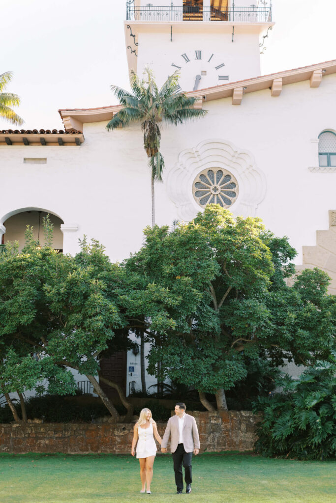 A couple holding hands walks on a grassy lawn in front of a white building with tall arched windows, a large clock, a palm tree, and lush green bushes. Copyright - Lisa Leanne Photography, California
