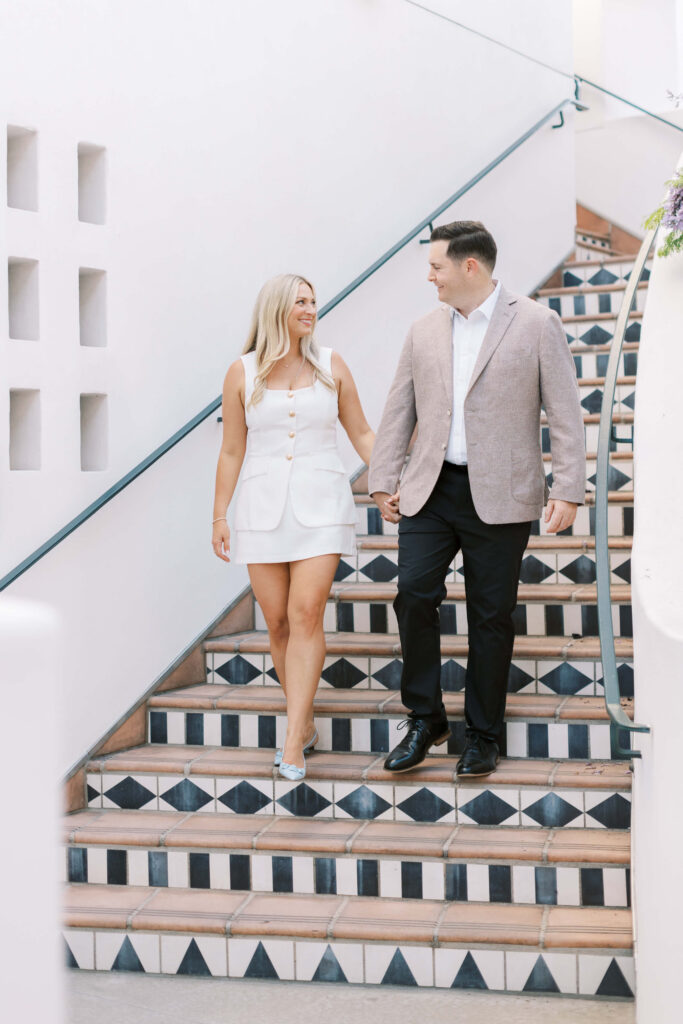 A couple walks down tiled stairs holding hands, smiling at each other. The woman wears a white dress and heels; the man wears a light blazer, white shirt, and black pants. The setting is bright with white walls. Copyright - Lisa Leanne Photography, California