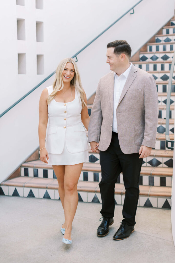 A smiling woman in a short white dress and a man in a beige blazer and black pants hold hands while walking indoors near a decorative tiled staircase. Copyright - Lisa Leanne Photography, California